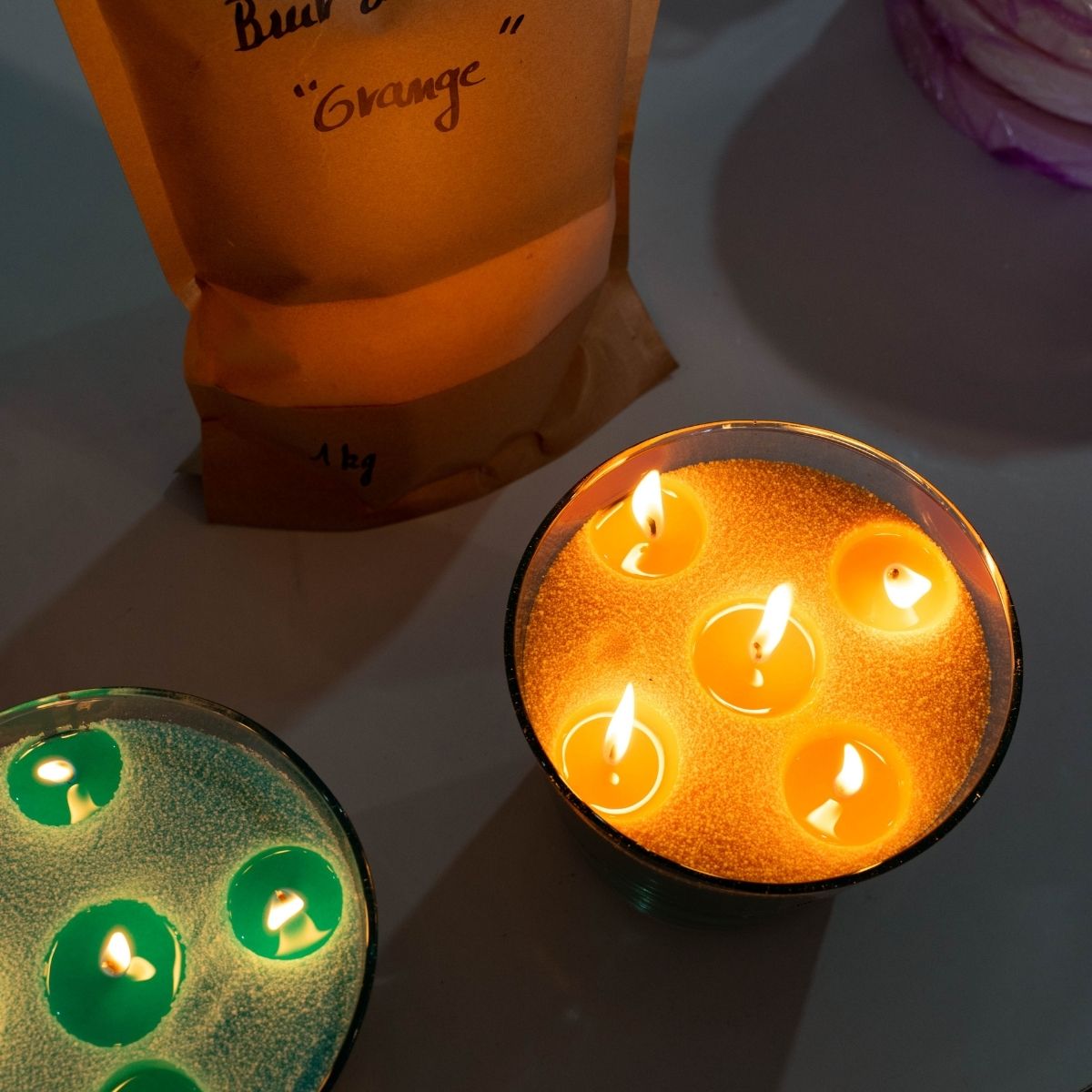 Two handcrafted, handmade glass vessels glow warmly — one filled with vibrant green sand wax, the other with warm orange sand wax. Both candles are ignited to illuminate elegant, serene spaces, showcasing Dary Living’s artisanal craftsmanship and rich, colorful wax for a refined home ambiance.